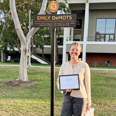 Emily DeMots, Employee of the Month for March 2025, stands under a sign bearing her name on campus.
