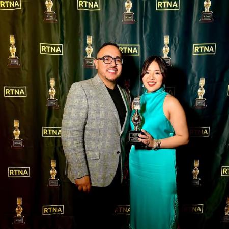 Two people stand in front of an RTNA step-and-repeat banner, with one person in dress holding a trophy shaped like a microphone