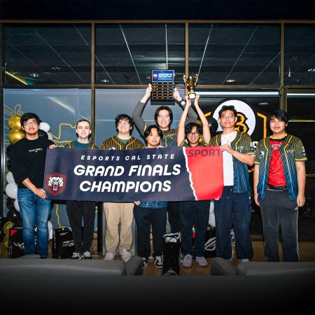 Group of eight people hold a "Grand Finals Champions" banner and two trophies at an esports event