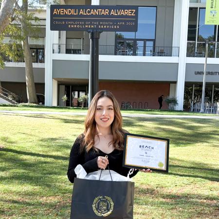 Employee of the Month Ayendili Alcantar Alvarez A person stands outside CSULB holding a framed award