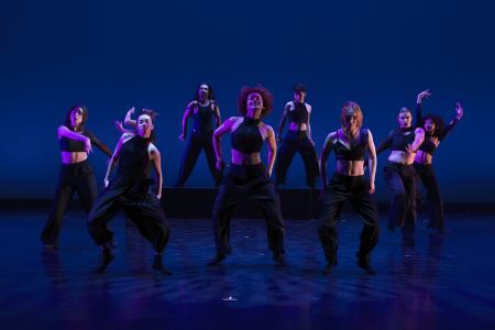 Eight dancers in black dance on a blue lit stage