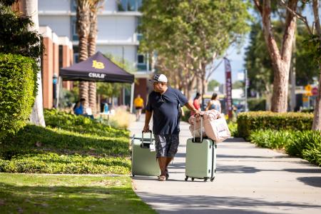 A person pulls two suitcases and bedding on a sidewalk near a CSULB tent