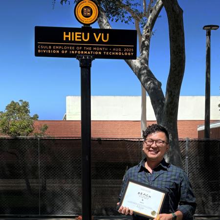Employee of the Month Hieu Vu A man smiles holding a certificate beneath a campus sign saying Hieu Vu
