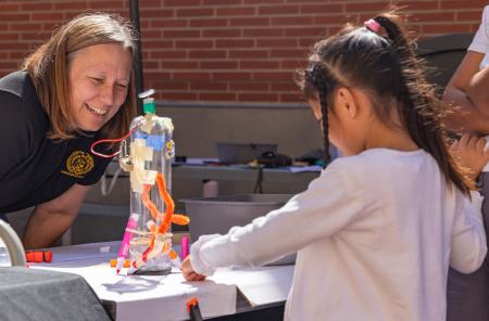 Educator watches a child assemble a hands-on science project at a table.
