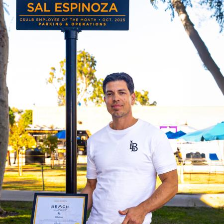 Employee of the Month Sal Espinoza stands under a sign bearing the same name while holding a framed certificate.