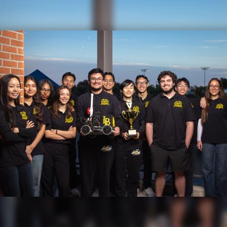 A group of CSULB students stands together as two hold a small robot and a trophy.