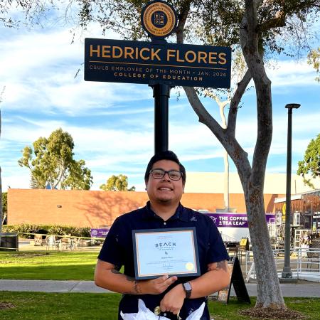 Employee of the Month Hedrick Flores stands on campus holding an award certificate beneath a named sign.