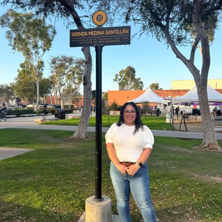 Employee of the Month Brenda Medina Santillán stands on campus beneath a sign displaying their name and recognition.
