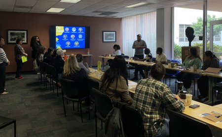 Community partners, 14 people, sit around a rectangle set up table with four people standing at the front of the room presenting from a screen