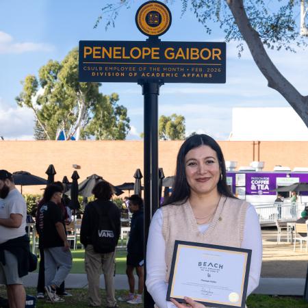 Person stands on campus holding a certificate beneath a sign displaying the name Penelope Gaibor.