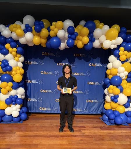 Picture of 1st place winner in biological and Agricultural Sciences, Amber Peek standing in front of a blue step and repeat surrounded by yellow, white, and blue balloons.