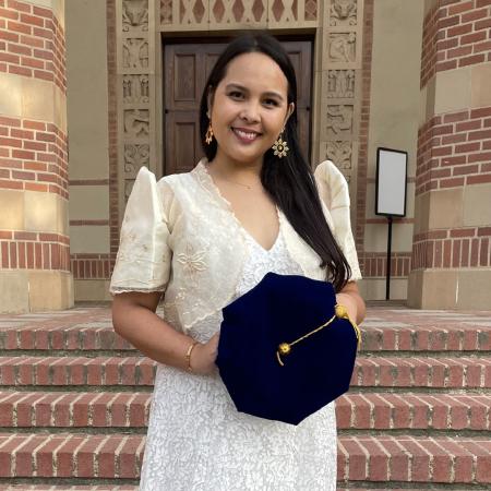 A person in formal attire holds a doctoral cap on steps outside a brick building