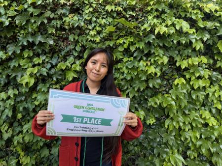A student standing in front of a wall with green leaves holding a sign