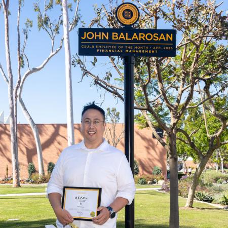 A person stands below a campus sign that says John Balarosan, Employee of the Month