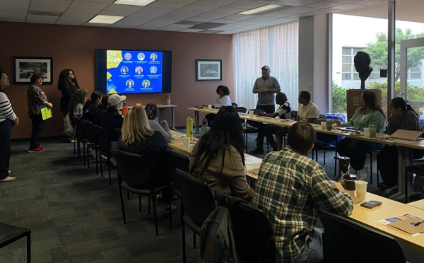 Community partners, 14 people, sit around a rectangle set up table with four people standing at the front of the room presenting from a screen