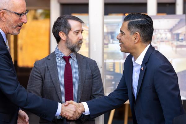 Two men shake hands with another man in the background