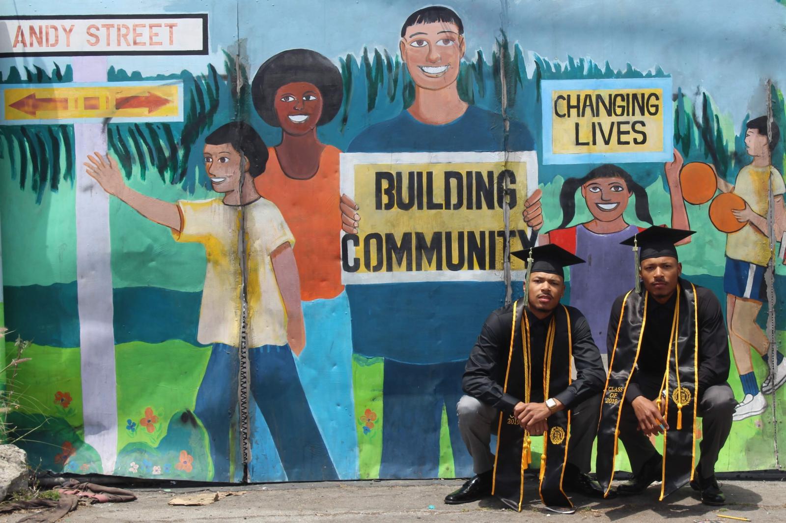 The Cooper twins, wearing CSULB caps and gowns, pose in front of a mural on on Andy Street where the grew up. The Cooper twins, wearing CSULB caps and gowns, pose in front of a mural on on Andy Street where the grew up.