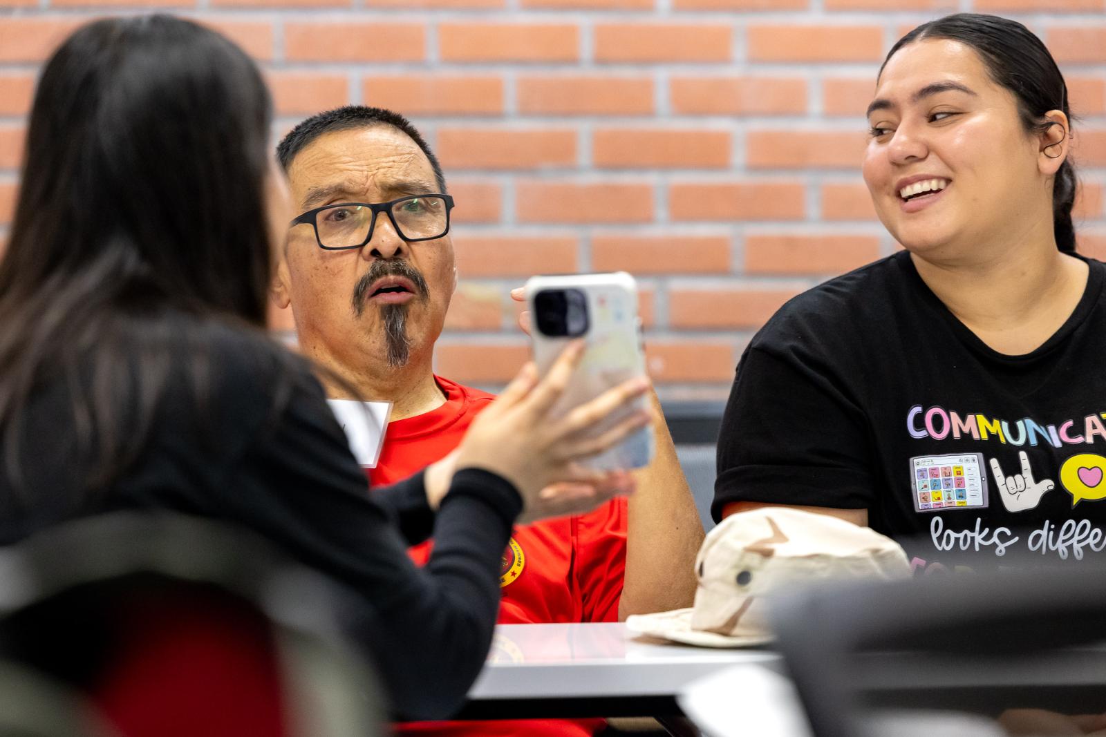 Three people talk at a table while one shows a phone