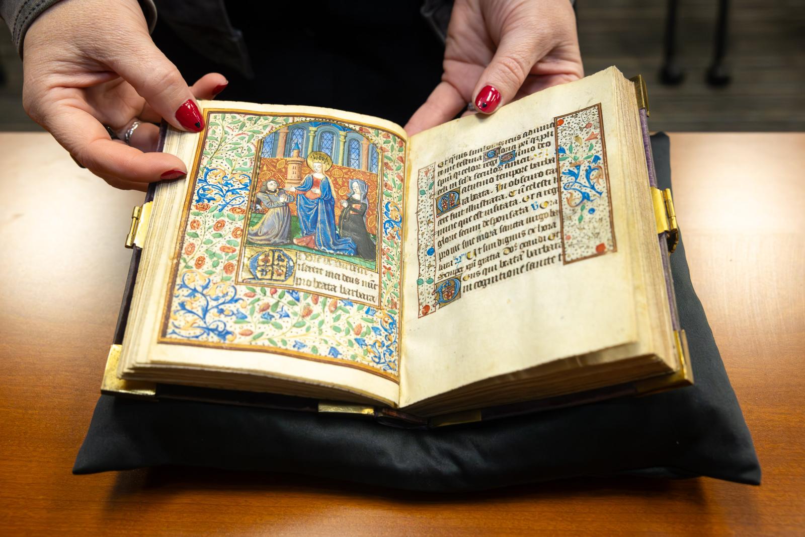 Hands hold open an illuminated manuscript with detailed illustrations and text on display.