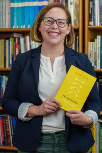 Dr. Fouratt is a white woman with chin length brown hair and glasses. She is wearing a blue blazer, white shirt, and green pants and is holding a yellow book.