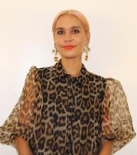 White woman, with blond hair, red lipstick, large earrings, and an animal print blouse
