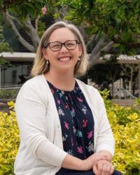 White woman with short brown hair, glasses, wearing a floral blouse and white sweater. She is sitting with her hand crossed on her lap. There are plants and trees in the background